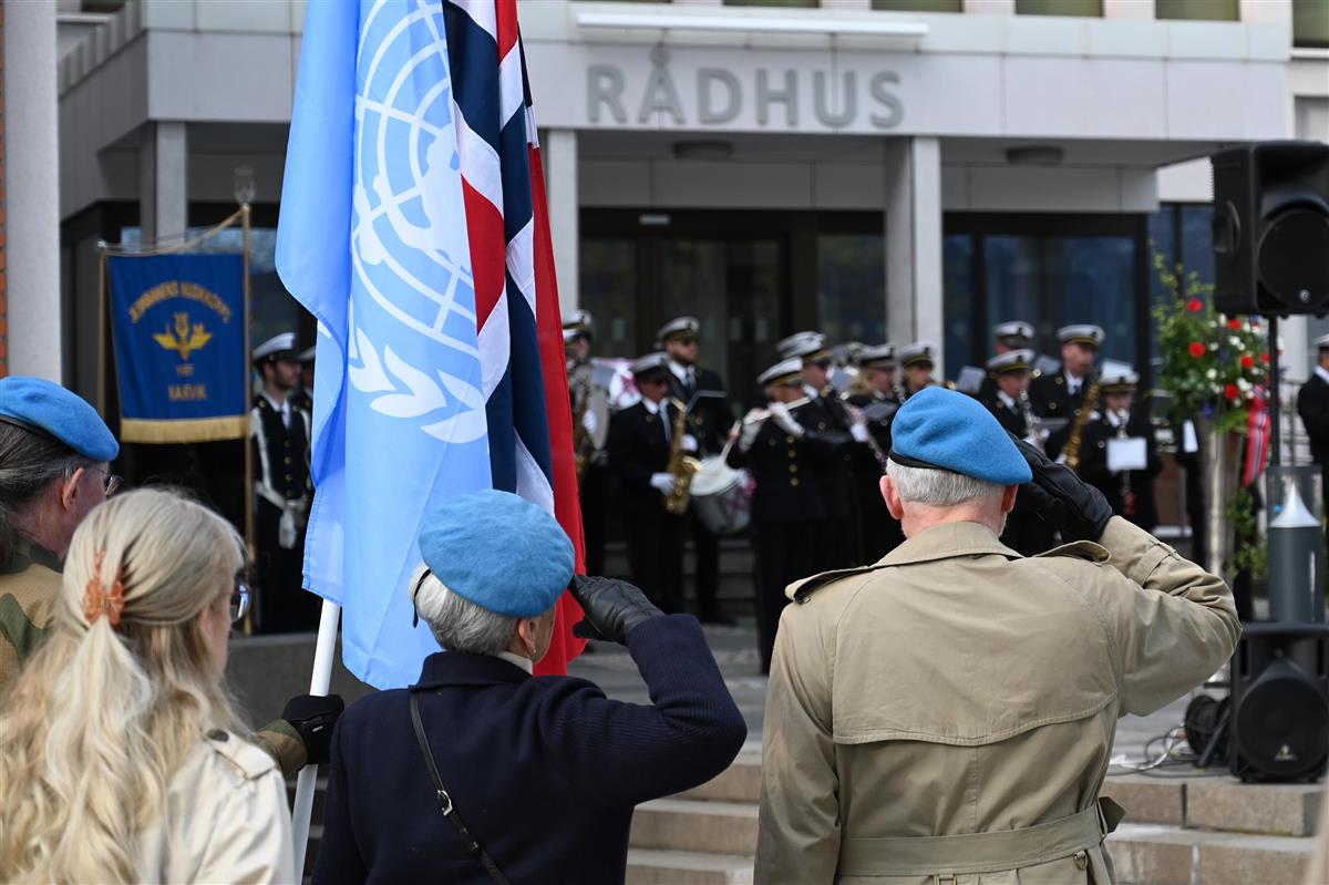 Militære veteraner med FN-bereter gjør honnør foran et rådhus, mens en fanebærer holder FN-flagget og det norske flagget foran et korps. - Klikk for stort bilde